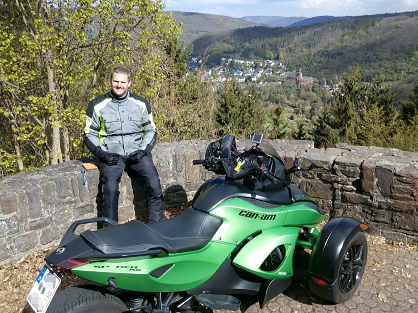 Mann sitzt in waldiger Landschaft auf Mauer, im Vordergrund ein Motorrad mit zwei Rädern vorne
