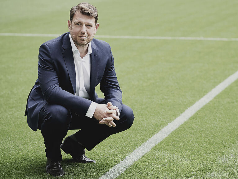 Phillip Laux im dunklen Anzug mit weißem Hemd, in der Hocke stehend auf Fußballrasen Phillip Laux im dunklen Anzug mit weißem Hemd, in der Hocke stehend auf Fußballrasen