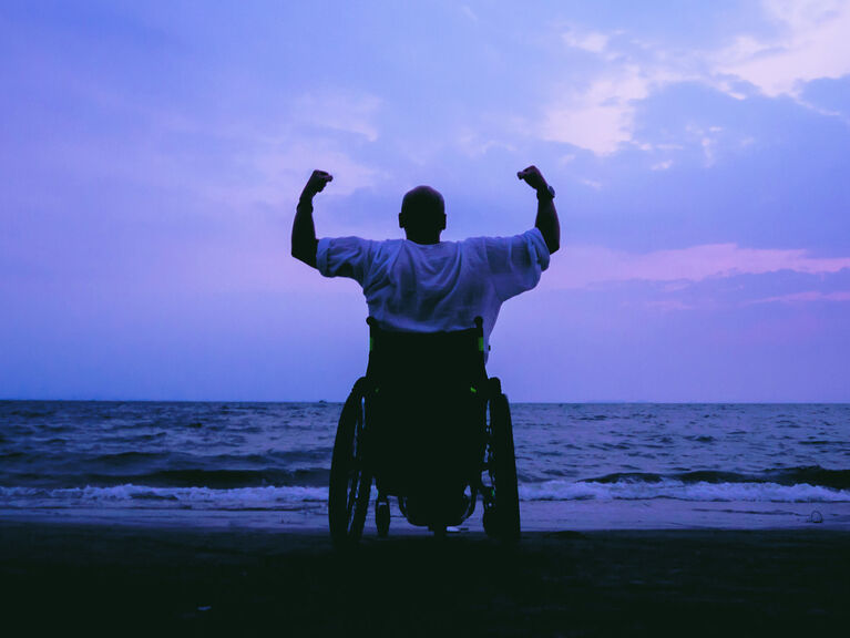 Rückenansicht eines Mannes im Rollstuhl, er hebt triumphierend beide Fäuste vor dem Meer in Abendstimmung Rückenansicht eines Mannes im Rollstuhl, er hebt triumphierend beide Fäuste vor dem Meer in Abendstimmung