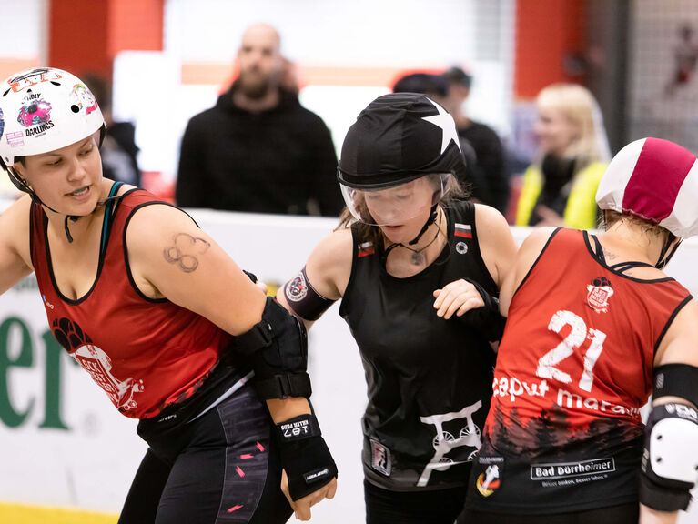 Drei Frauen in Sportkleidung und Schutzhelmen rempeln sich gegenseitig im Rahmen eines sportlichen Wettkampfes an