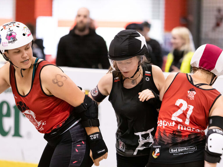 Drei Frauen in Sportkleidung und Schutzhelmen rempeln sich gegenseitig im Rahmen eines sportlichen Wettkampfes an Drei Frauen in Sportkleidung und Schutzhelmen rempeln sich gegenseitig im Rahmen eines sportlichen Wettkampfes an