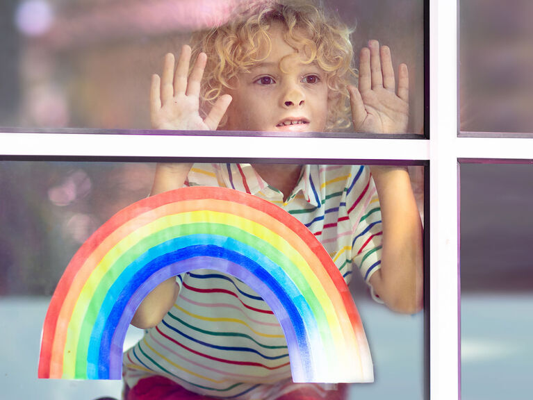 Kleiner Junge presst Hände und Gesicht traurig gegen eine Fensterscheibe, in der ein Regenbogen aus Papier klebt