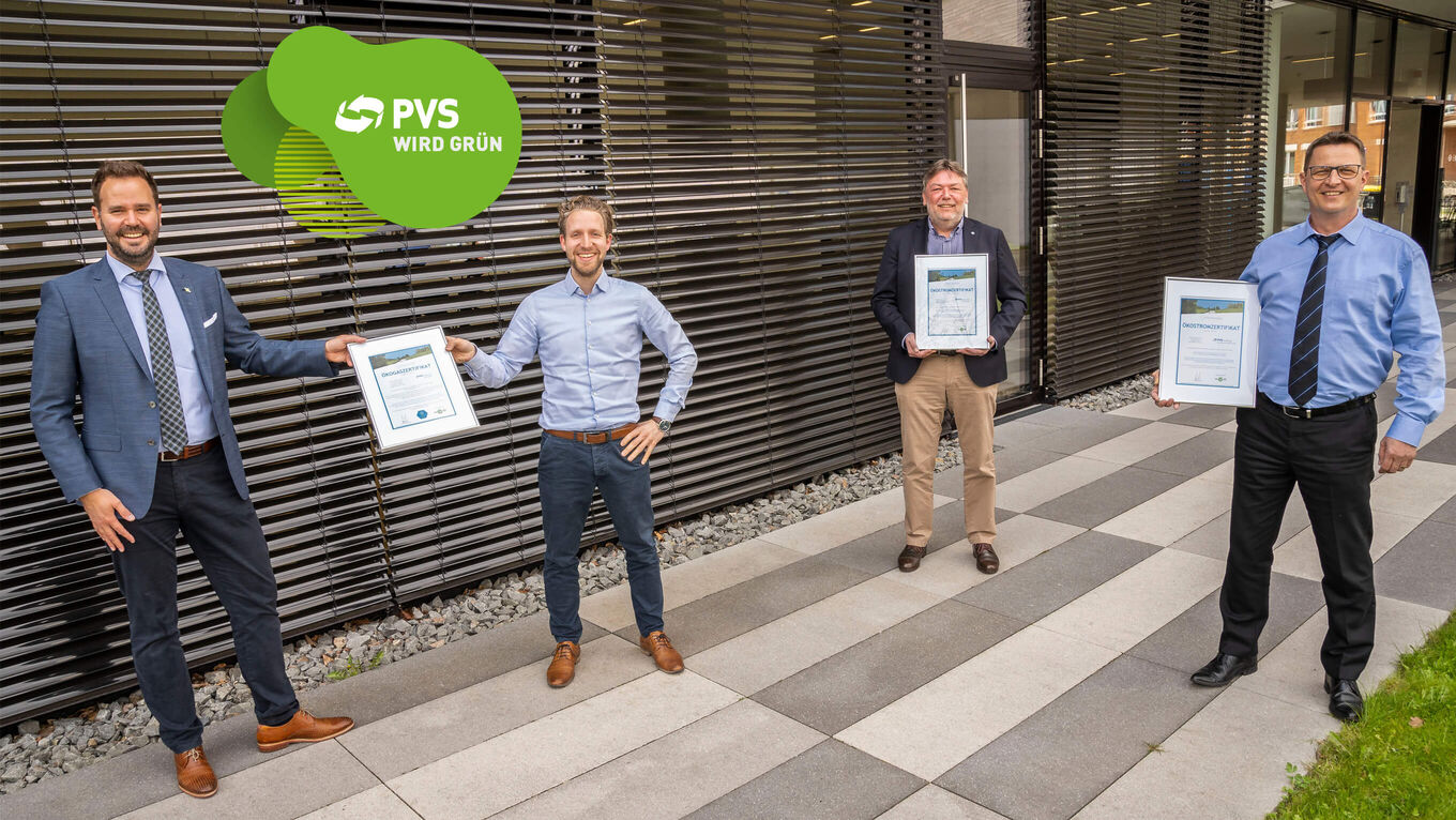 Vier Männer stehen mit Abstand voneinander vor einem Bürogebäude und halten Zertifikate hoch. Oben links im Bild: Logo "PVS wird grün"