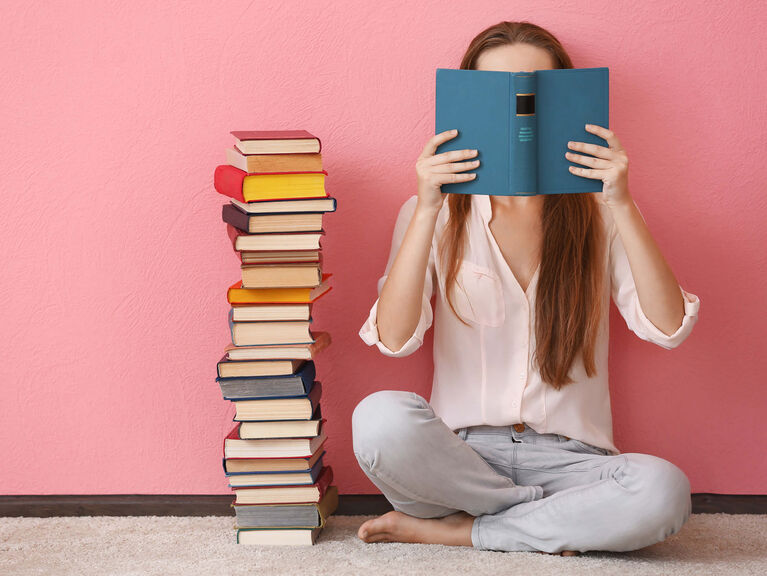 Frau sitzt neben einem hohen Bücherstapel im Schneidersitz vor einer rosafarbenen Wand und hält ein offenes Buch vor ihr Gesicht Frau sitzt neben einem hohen Bücherstapel im Schneidersitz vor einer rosafarbenen Wand und hält ein offenes Buch vor ihr Gesicht