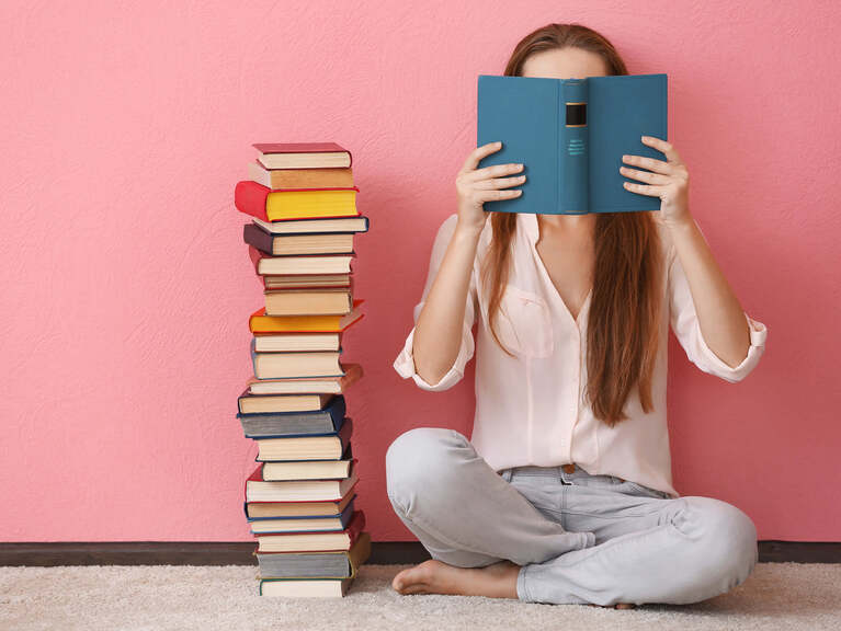 Frau sitzt neben einem hohen Bücherstapel im Schneidersitz vor einer rosafarbenen Wand und hält ein offenes Buch vor ihr Gesicht Frau sitzt neben einem hohen Bücherstapel im Schneidersitz vor einer rosafarbenen Wand und hält ein offenes Buch vor ihr Gesicht