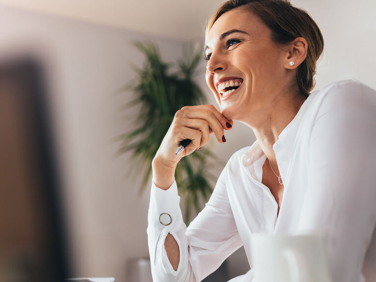 Lachende Frau in weißer Bluse sitzt mit den Ellbogen aufgestützt am Schreibtisch nebst Laptop und Kaffeetasse im Vordergrund