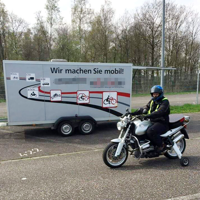 Mann sitzt auf Motorrad mit Stützrädern auf einem asphaltierten Platz, im Hintergrung steht ein Anhänger mit der Aufschrift "Wir machen Sie mobil"