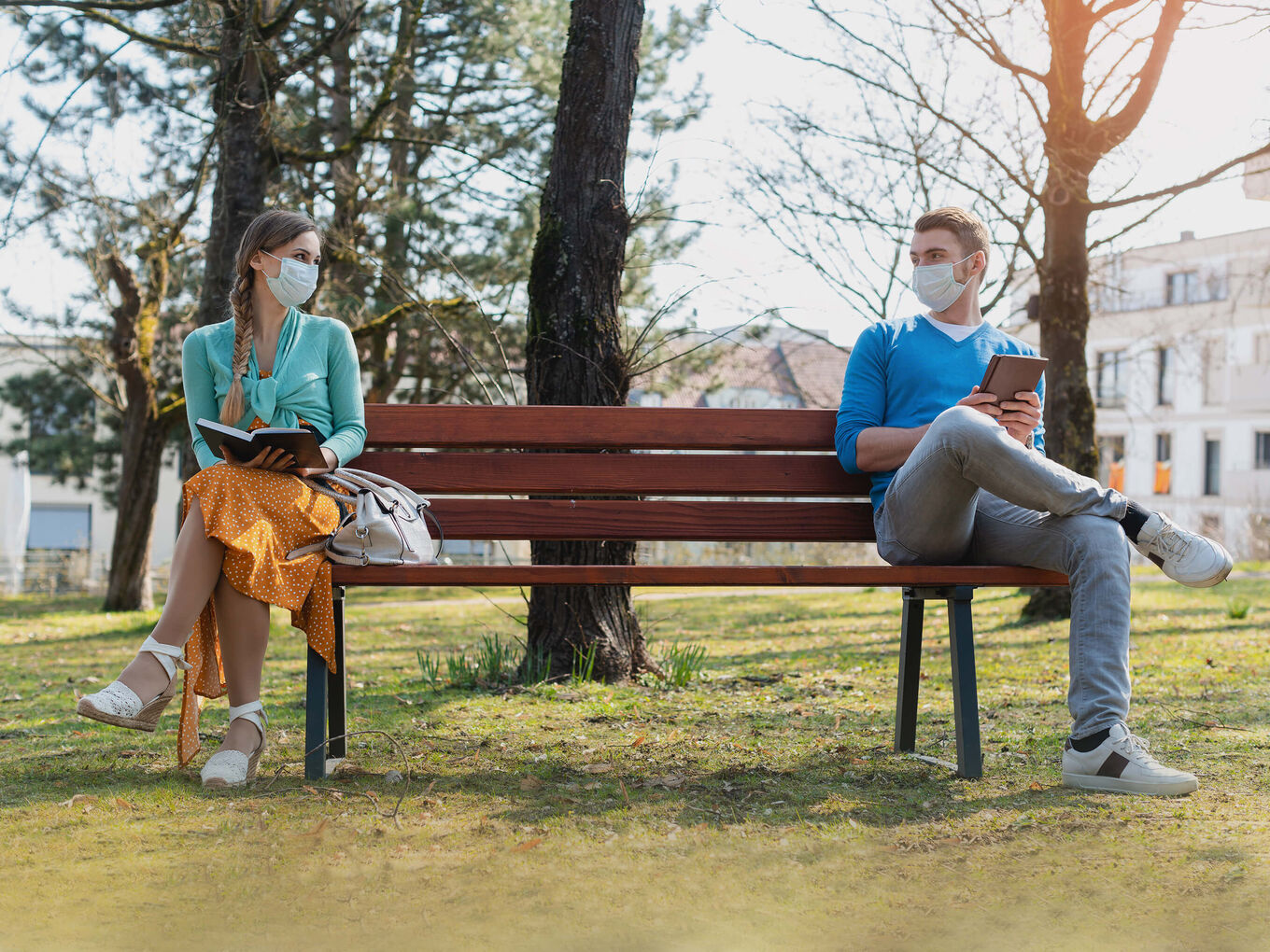 Junge Frau und junger Mann, jeweils mit OP-Maske, sitzen mit geöffneten Büchern in der Hand auf einer Bank im Park und schauen sich freundlich an