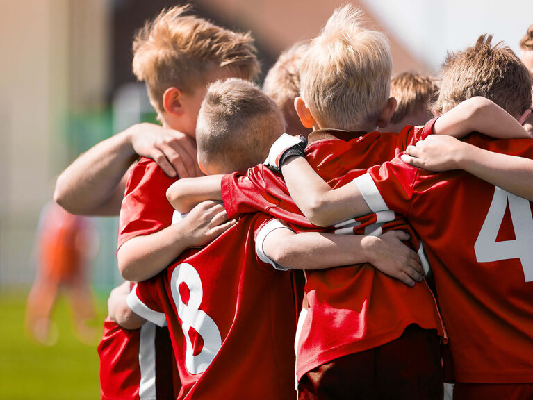 Rückenansicht einer Gruppe von Jungs im Grundschulalter, sich im Kreis stehend umarmend, rote Fußballtrikots tragend