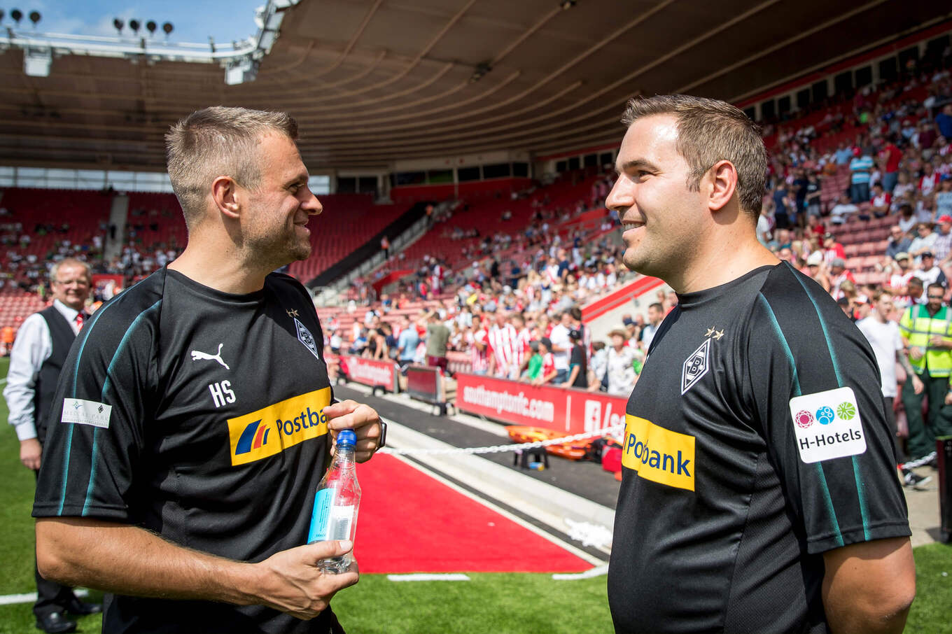 Zwei Männer in schwarzen Sporttrikots unterhalten sich lächelnd, im Hintergrund Zuschauerränge eines Fußballstadions