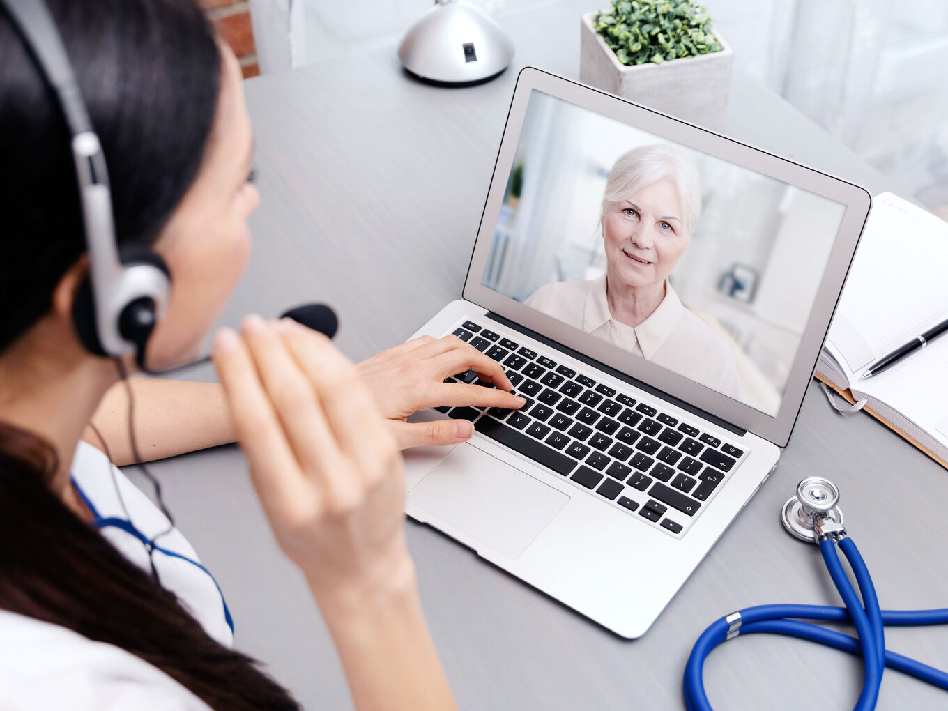 Junge Ärztin mit Headset sitzt am Schreibtisch und spricht mit Patientin über einen Laptopscreen