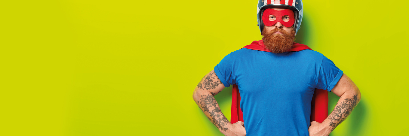 Direkter, ernsthaft-humorvoller Blick eines bärtigen, tätowierten Mannes in Superheldenpose (geballte Fäuste in den Hüften), mit Cape, Helm und Augenmaske