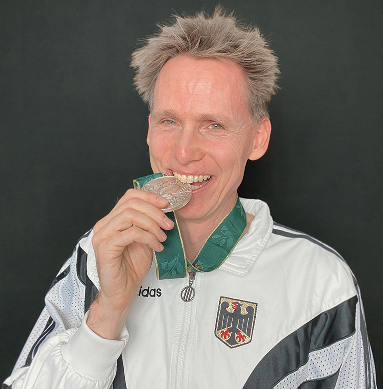 Frank Busemann, eine weiße Trainingsjacke mit Bundesadler-Emblem tragend, beißt lächelnd in seine olympische Goldmedaille