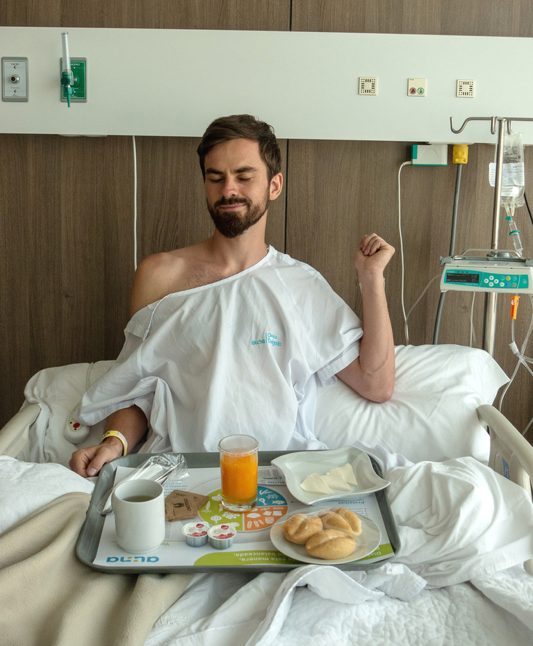 Patrick Arendt sitzt in Krankenhaushemd in einem Krankenhausbett und freut sich über ein volles Essenstablett auf seinem Schoß