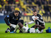 Zwei Männer betreuen einen am Boden liegenden Fußballer medizinisch auf einem Fußballrasen, im Hintergrund Publikumsränge eines Stadions