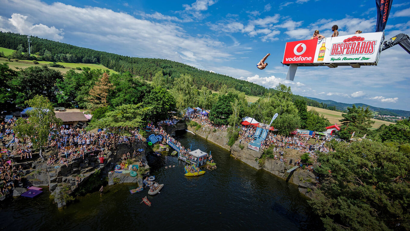 Luftbild: Turmspringer springt von Sprungturm, Wälder, Felder und ein See mit Publikum am Rand sind im Hintergrund sichtbar