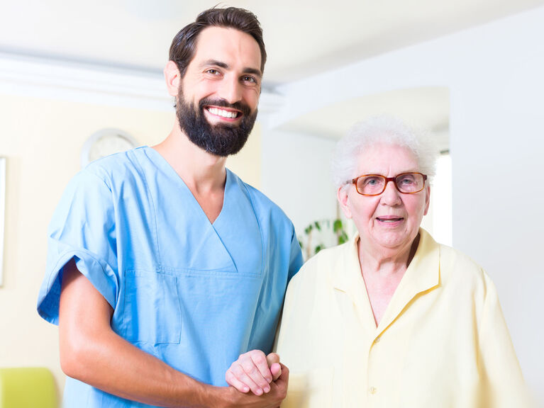 Junger, sympathisch lächelnder Pfleger im blauen Kasak hält stützend die Hand einer fröhlichen Seniorin