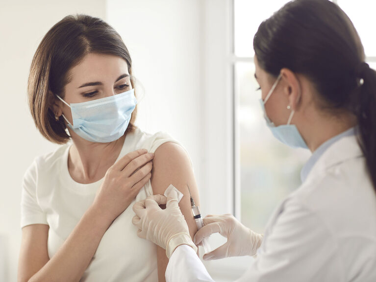 Frau mit Mund-Masen-Schutz bekommt Impfung in den Oberarm von Ärztin in Rückenansicht