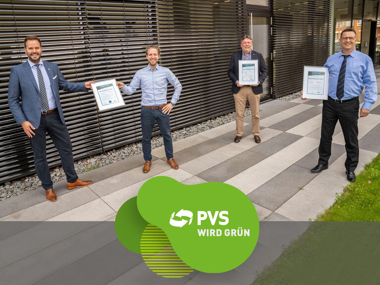 Vier Männer stehen mit Abstand voneinander vor einem Bürogebäude und halten Zertifikate hoch. Mittig im Bild: Logo "PVS wird grün"