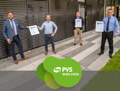 Vier Männer stehen mit Abstand voneinander vor einem Bürogebäude und halten Zertifikate hoch. Mittig im Bild: Logo "PVS wird grün"
