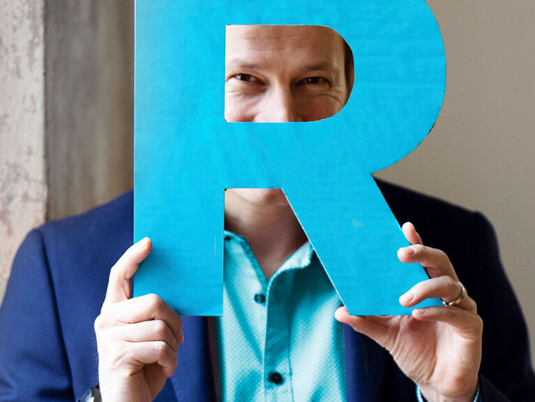 Tim Niedernolte hält ein großes, cyanfarbenes "R" aus Pappe hoch und schaut durch die runde Aussparung des Buchstaben Tim Niedernolte hält ein großes, cyanfarbenes "R" aus Pappe hoch und schaut durch die runde Aussparung des Buchstaben