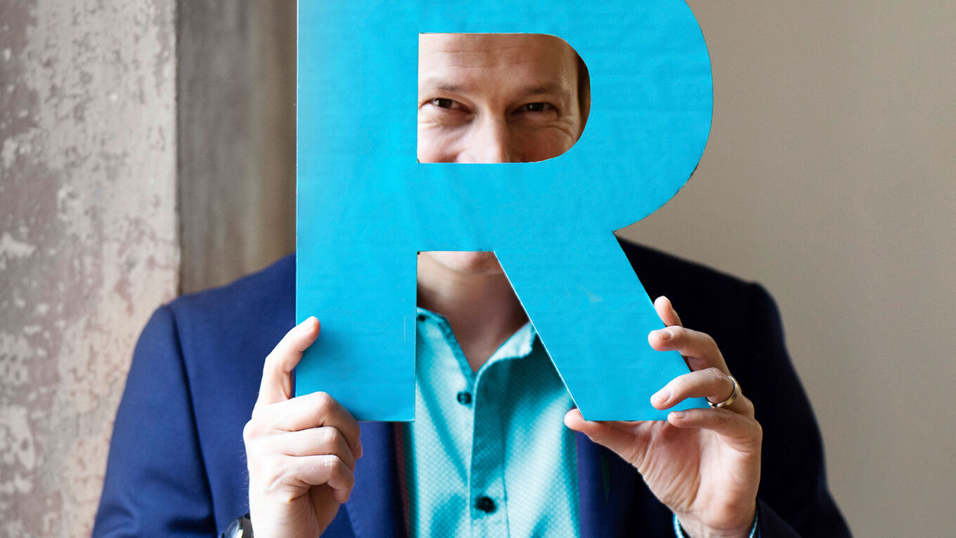 Tim Niedernolte hält ein großes, cyanfarbenes "R" aus Pappe hoch und schaut durch die runde Aussparung des Buchstaben