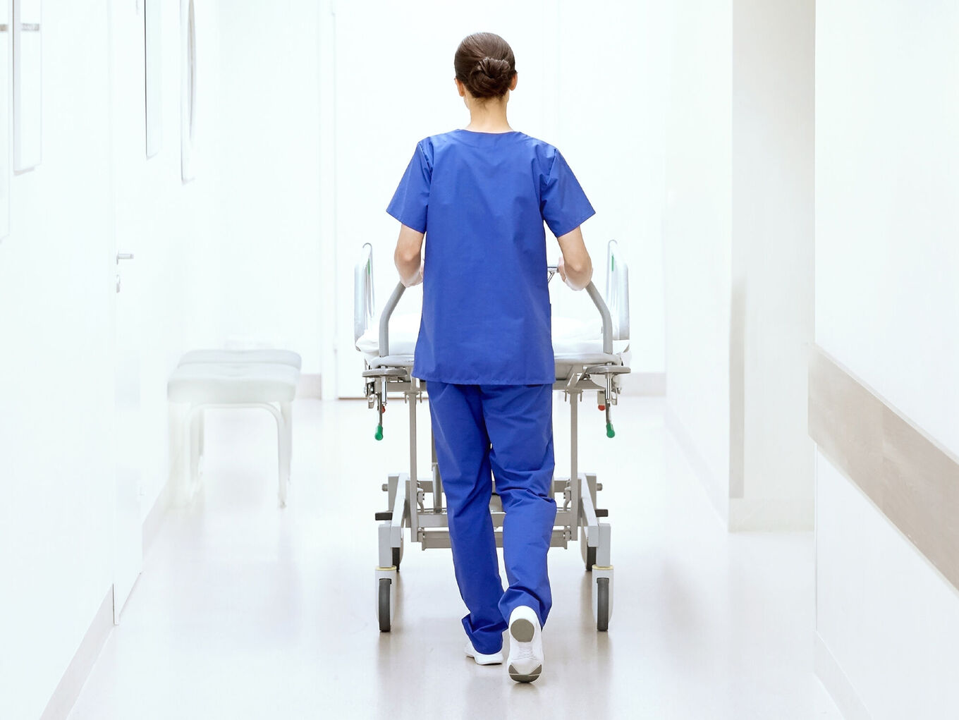 Rückansicht einer Pflegerin in blauer OP-Kleidung, ein Bett über einen Krankenhausgang schiebend