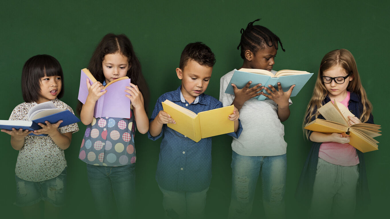 Fünf ethnisch diverse Kinder stehen nebeneinander lesend vor einer dunkelgrünen Tafel