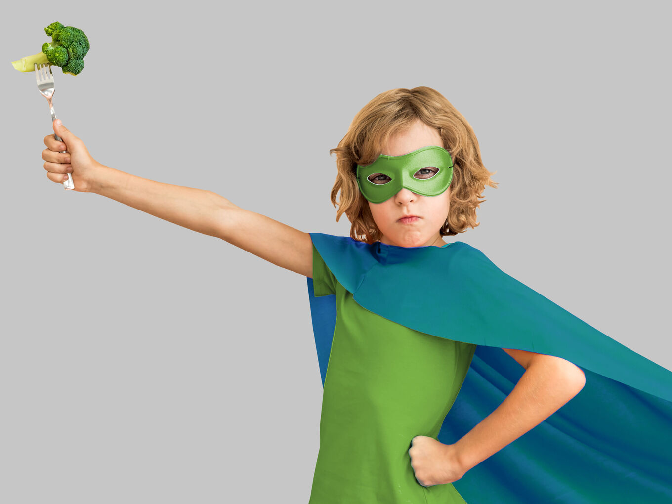 Kind in Superheldenmontur hält Gabel mit Brokkoli in die Luft