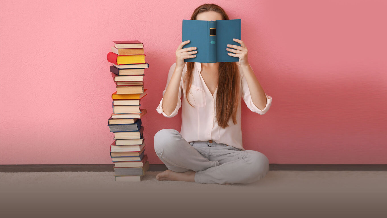 Frau sitzt neben einem hohen Bücherstapel im Schneidersitz vor einer rosafarbenen Wand und hält ein offenes Buch vor ihr Gesicht