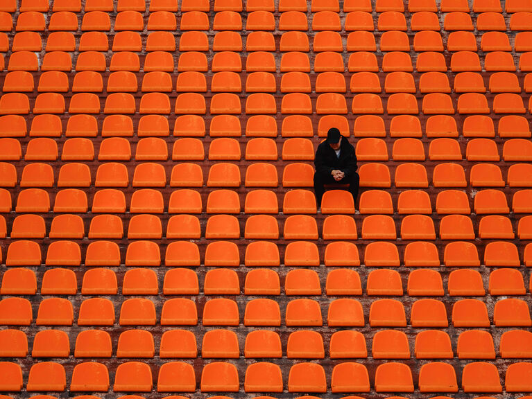 Viele, leere Stadionsitzplätze aus orangefarbenem Plastik, ein Platz ist besetzt von einer traurigen Gestalt in schwarz Viele, leere Stadionsitzplätze aus orangefarbenem Plastik, ein Platz ist besetzt von einer traurigen Gestalt in schwarz